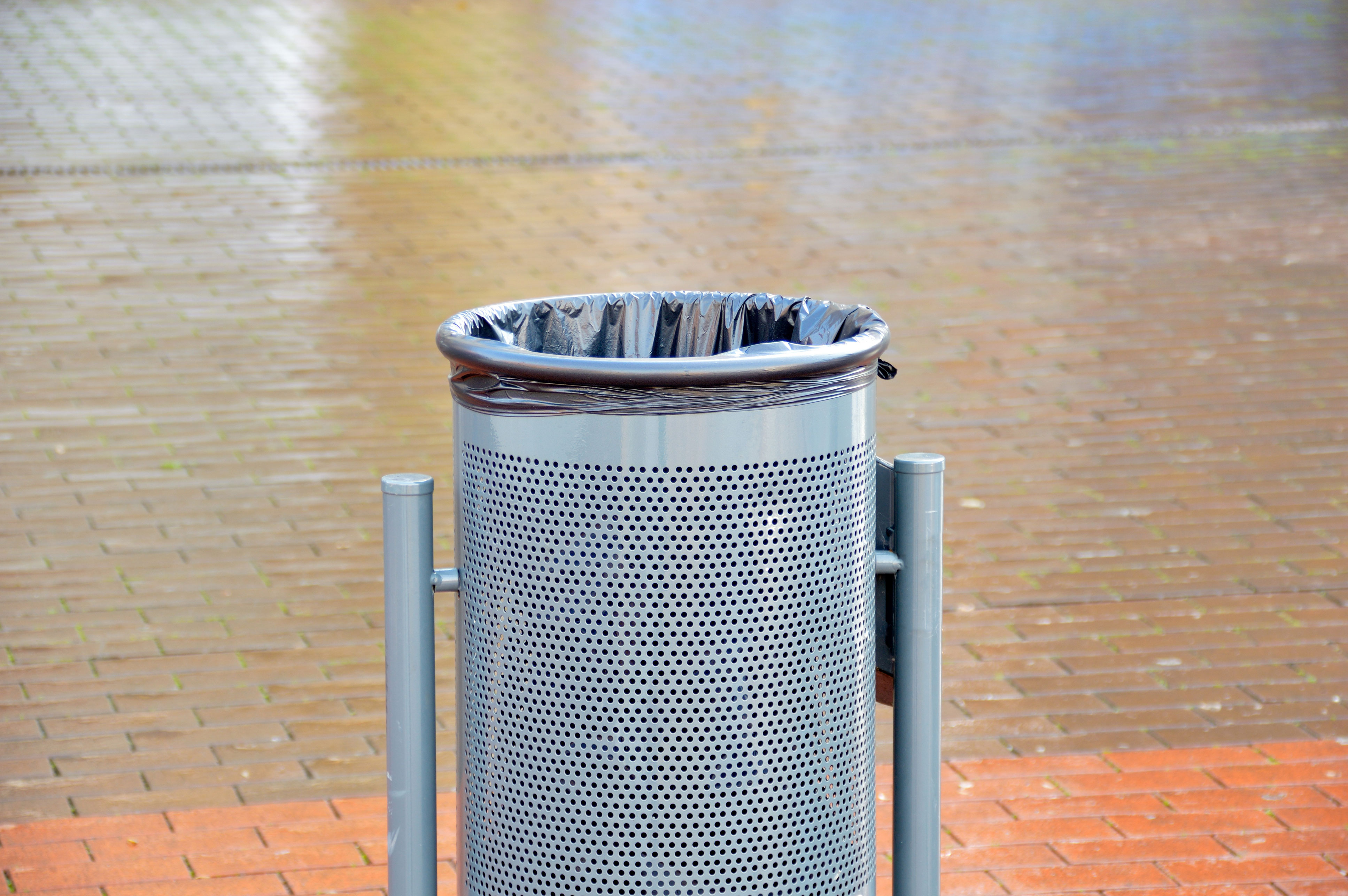 Papelera urbana cilíndrica de acero perforado en color gris, con estructura metálica de soporte y abertura superior para depositar residuos, instalada en una zona peatonal pavimentada