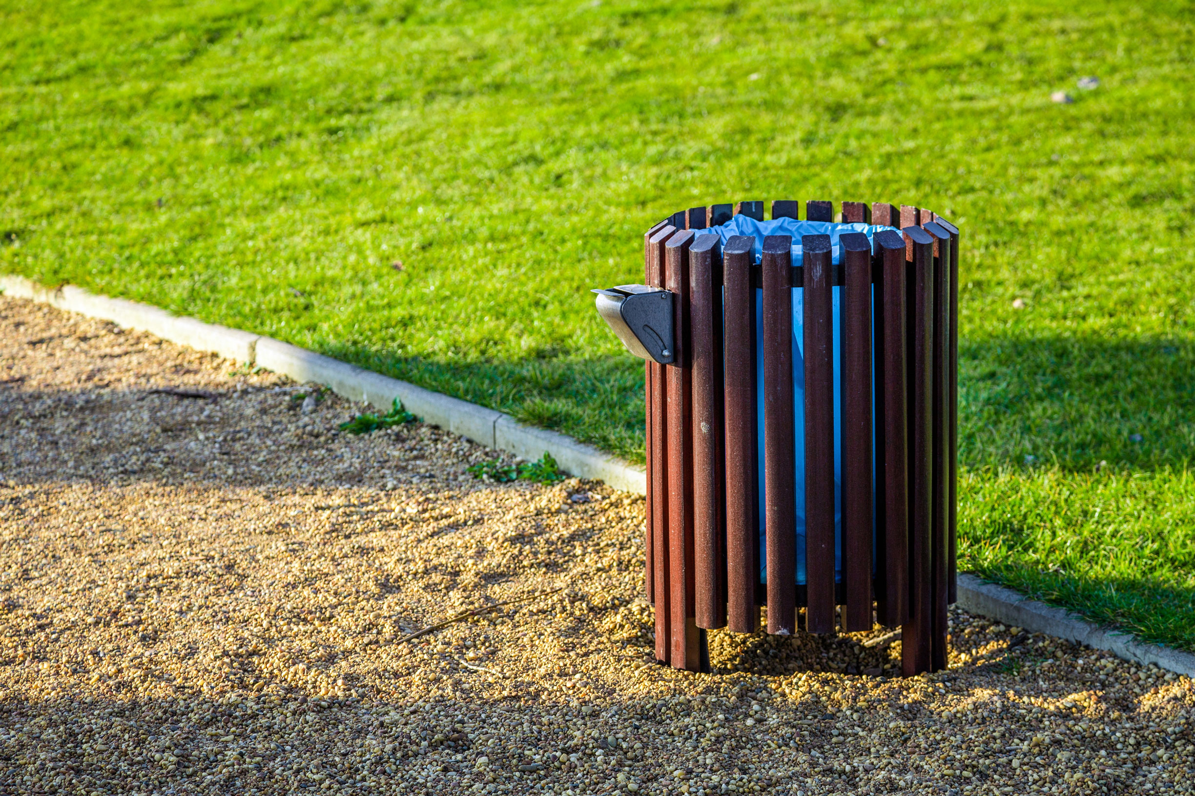 Papelera urbana circular de madera con lamas verticales marrones, estructura metálica y depósito interior, instalada junto a una zona ajardinada de un parque público para la recogida de residuos