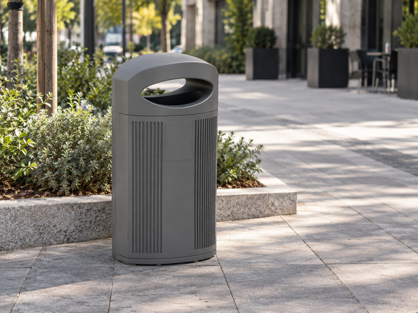 Papelera urbana de plástico gris con diseño moderno, ranuras verticales y abertura ovalada, instalada sobre el pavimento de una plaza con zonas ajardinadas