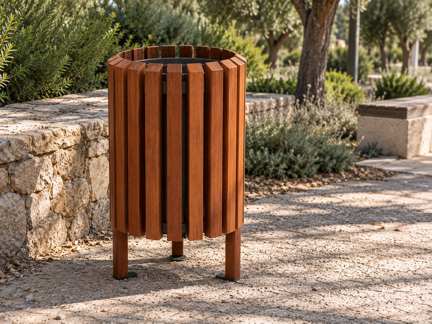 Papelera urbana de madera con diseño circular y lamas verticales, instalada en un parque rústico con vegetación mediterránea y muro de piedra natural