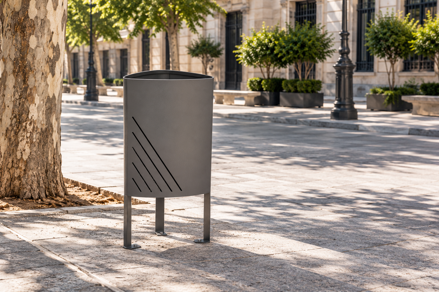Papelera urbana de acero modelo Élite en color gris antracita, instalada en una plaza pública con pavimento de piedra y zonas ajardinadas